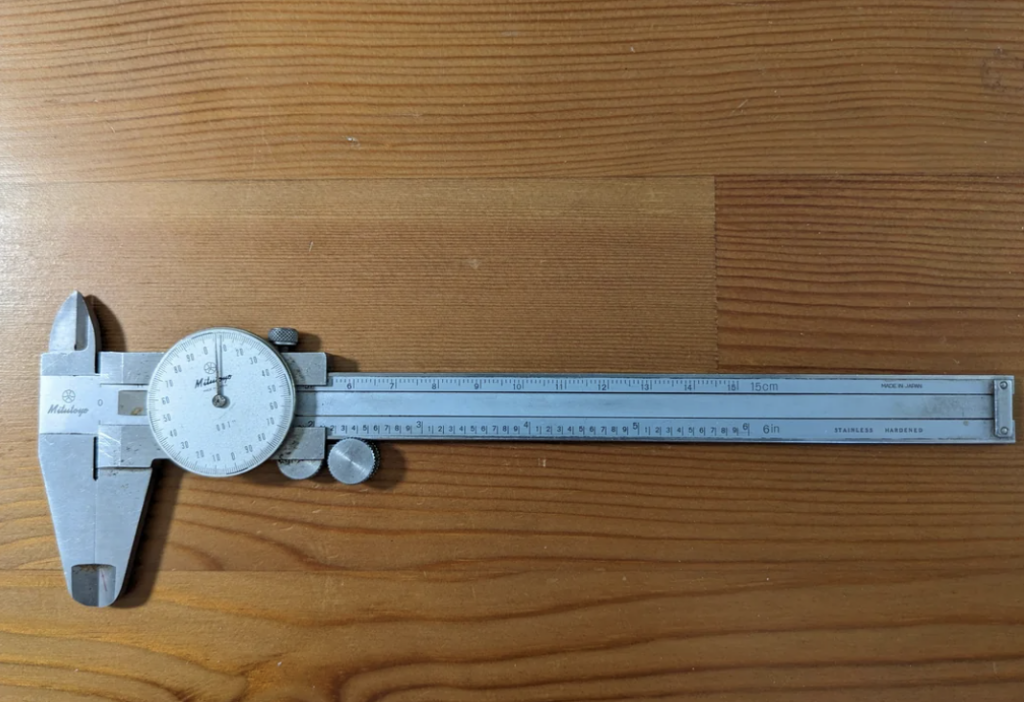 A silver dial caliper with a circular gauge and measurement scale displayed on a wooden surface. The caliper is marked with increments in both inches and centimeters, and the dial shows small, precise increments for accurate measuring.