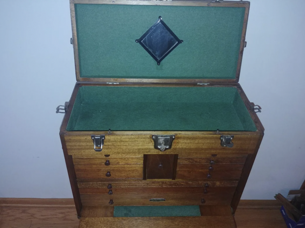 Open wooden tool chest with a green felt lining, featuring multiple drawers and metal latches. The lid has a diamond-shaped black detail on the felt interior. The chest is situated against a white wall on a wooden floor.