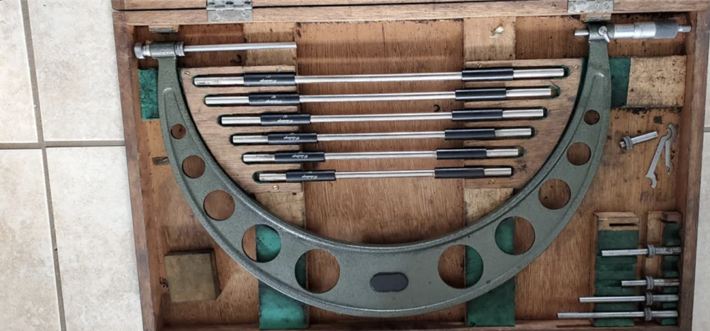 A wooden case containing a large metal arc-shaped tool with circular cutouts, alongside a set of rods and various small metal components, all neatly arranged in compartments. The case is open on a tiled floor.