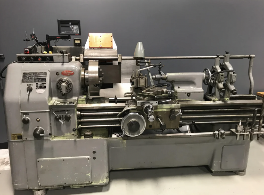 A large metal lathe machine with various levers, knobs, and controls situated in a workshop. The machine has a digital display monitor and a gray body with areas of exposed metal.