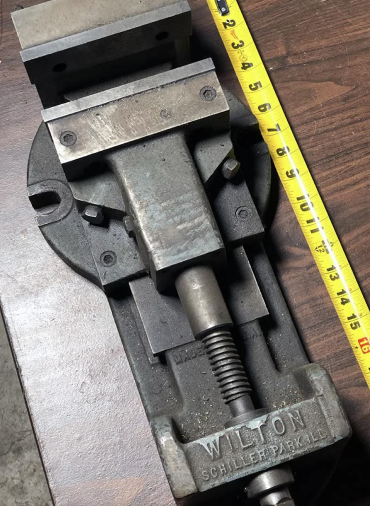 A metal Wilton vise on a wooden surface with a yellow measuring tape extended along its length. The vise has a screw mechanism and metal jaws, with the brand name "Wilton" and additional text visible.