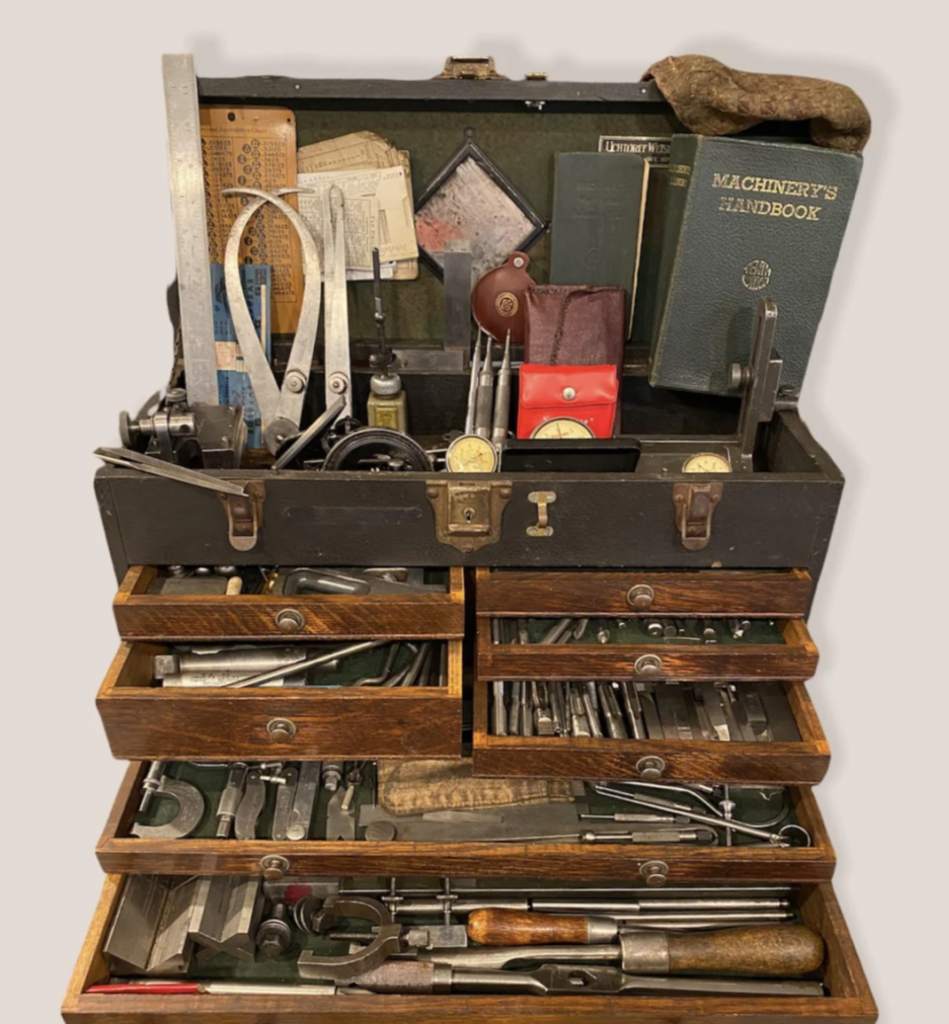 A vintage toolbox with multiple open wooden drawers containing various metal tools, including calipers, wrenches, and gauges. On top are a Machinery's Handbook, a brush, and additional items.