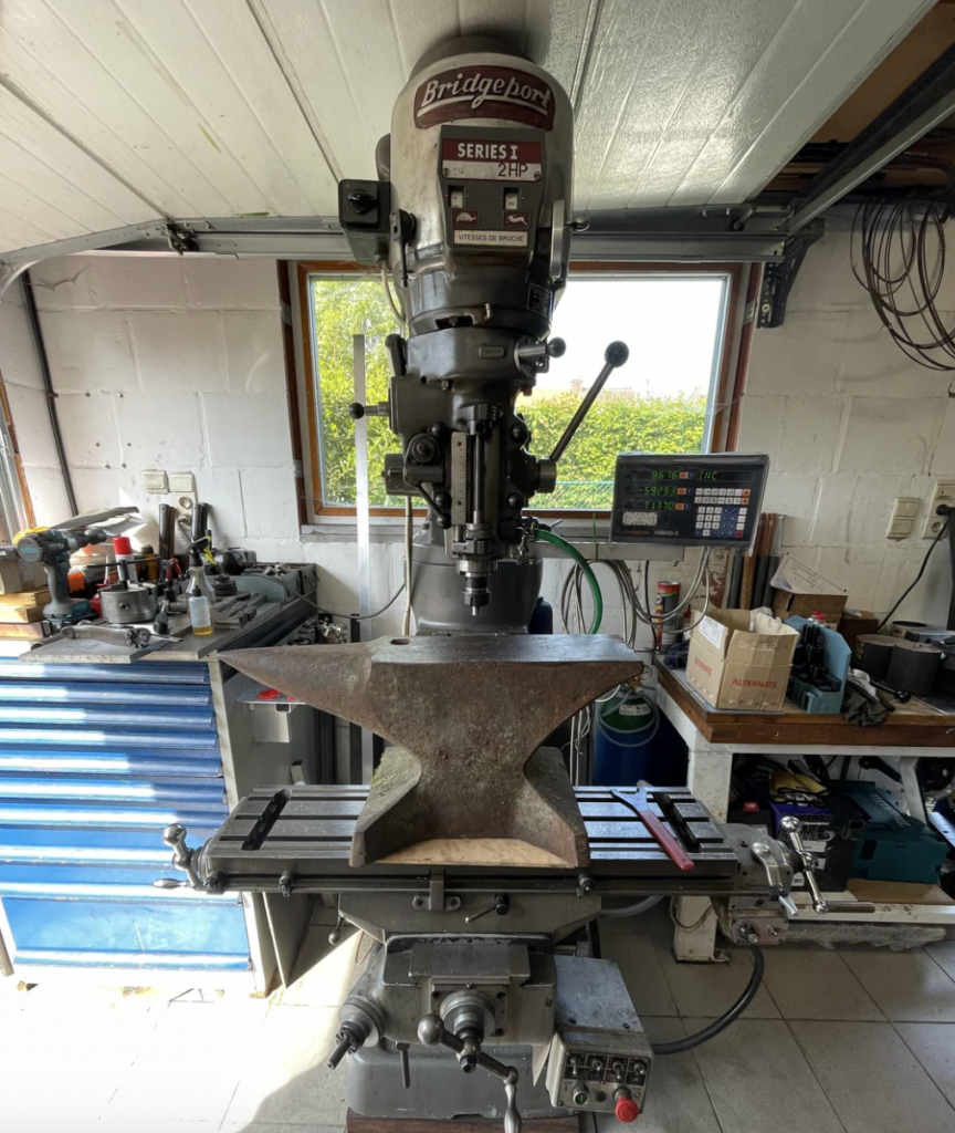 A Bridgeport milling machine in a workshop holds a large metal anvil on its table. There's a digital display on the side, with tools and boxes visible in the background on shelves and a workbench.