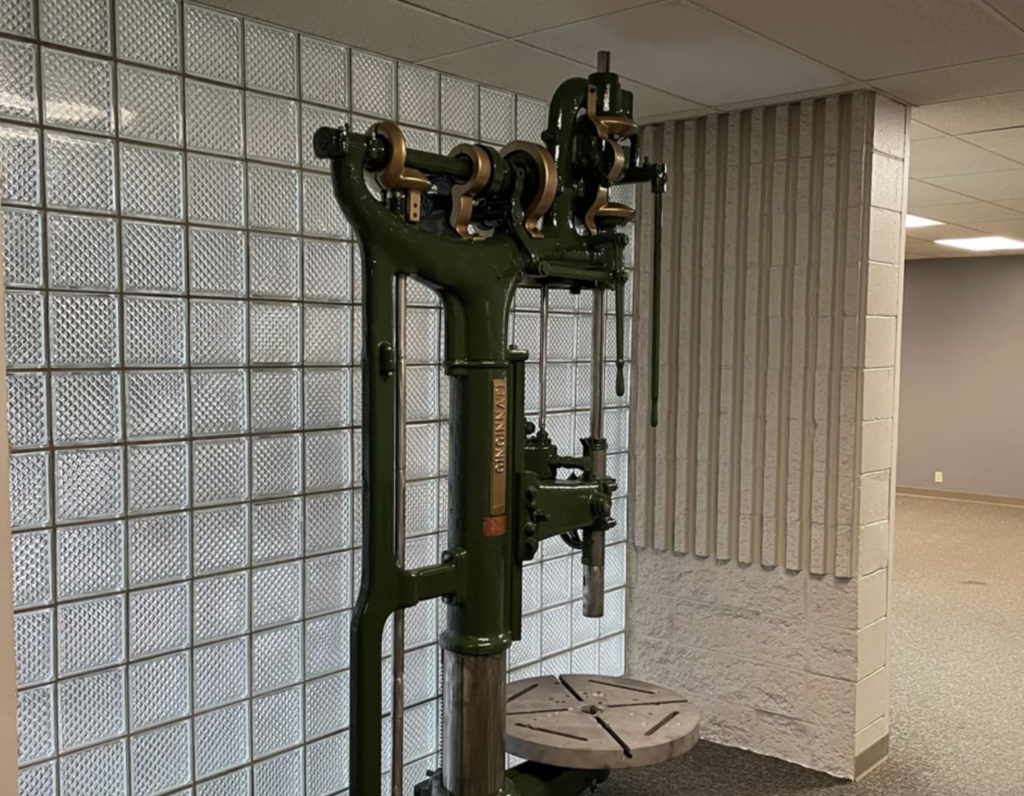 A vintage industrial green drill press with metal and wooden components is set against a backdrop of glass block walls and vertical grooves. The drill has a flat circular base and multiple gears and levers.