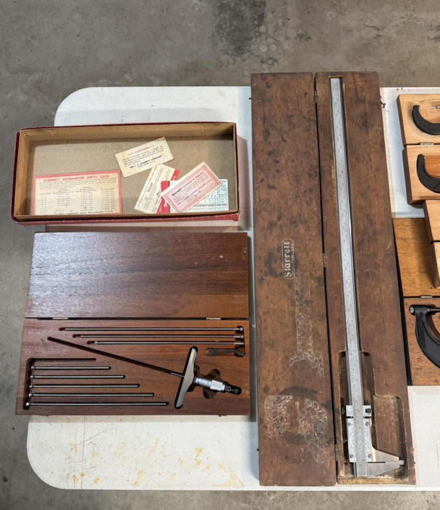 A wooden box containing metal alignment tools and a T-shaped instrument is open on a table. Nearby, a long measuring tool is in another wooden box. Some papers and a cardboard lid with calibration tables are also visible.