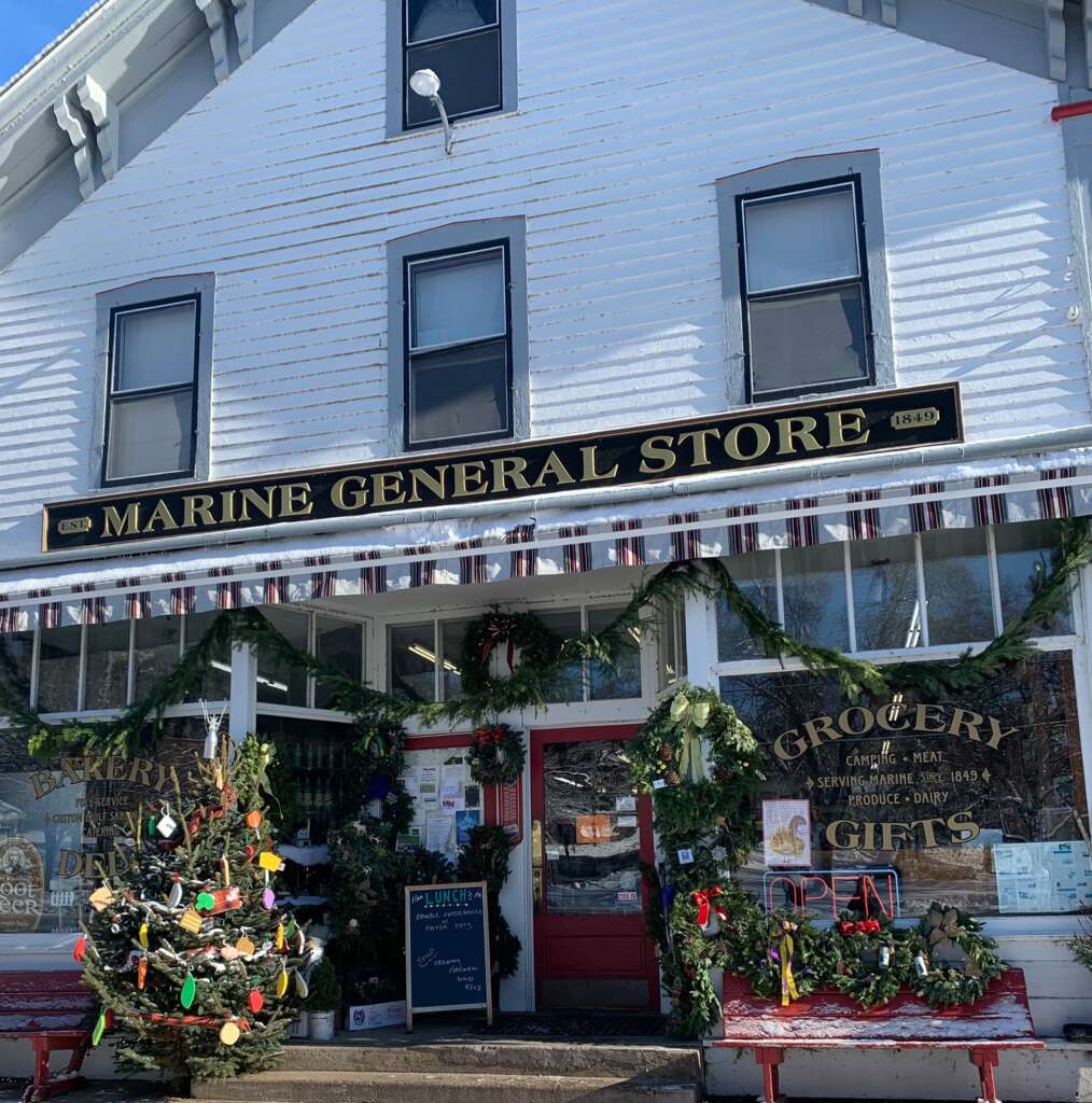 A charming white building houses the Marine General Store. The entrance is adorned with festive garlands and a small decorated Christmas tree. Signs in the window offer bakery goods and groceries.