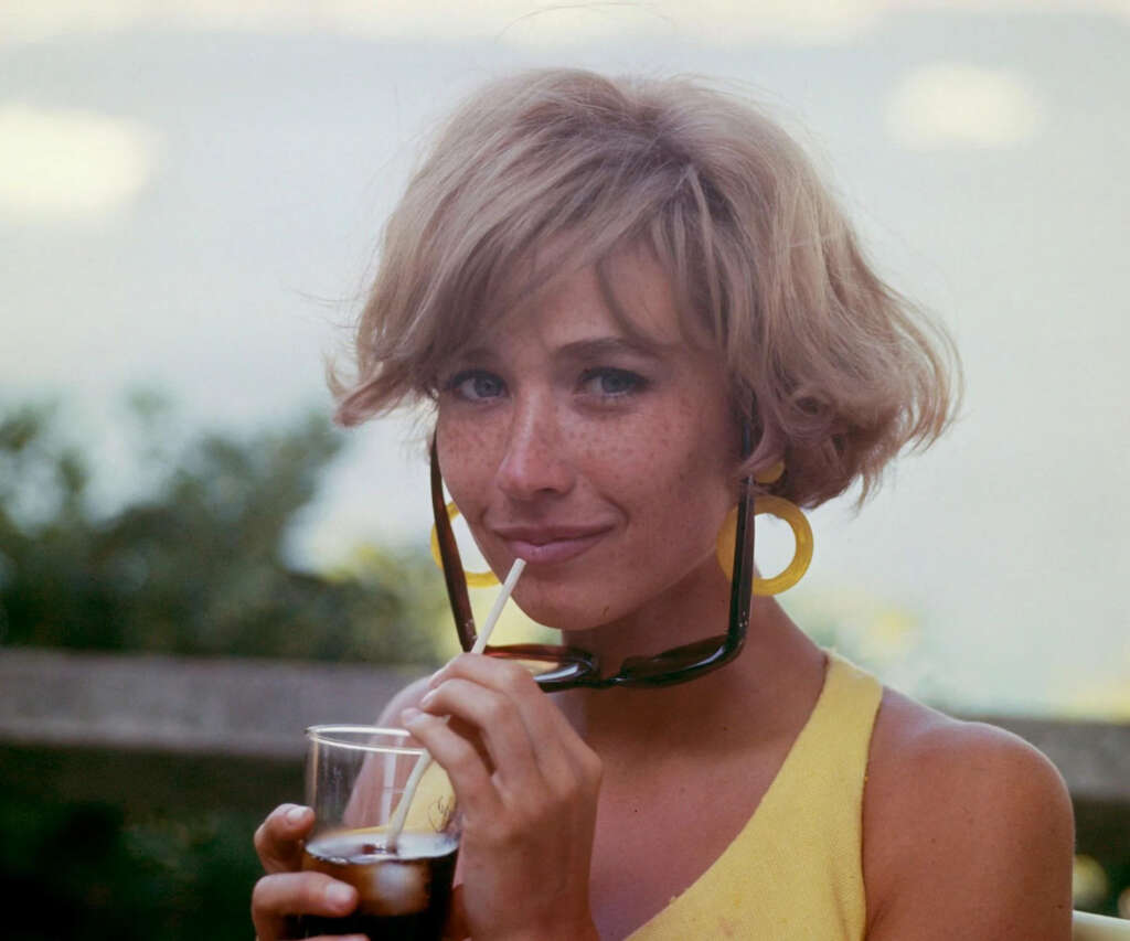 A person with short blonde hair and large hoop earrings sips a drink with a straw. They are wearing a yellow top and sunglasses are tucked into their shirt. The background is blurred with greenery and water.