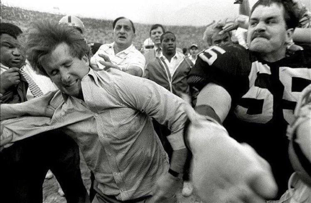 A black and white photo shows a chaotic scene during a football game. A man in a shirt is being punched by a player wearing a number 55 jersey. A crowd of people surrounds them, capturing a moment of tension and conflict.