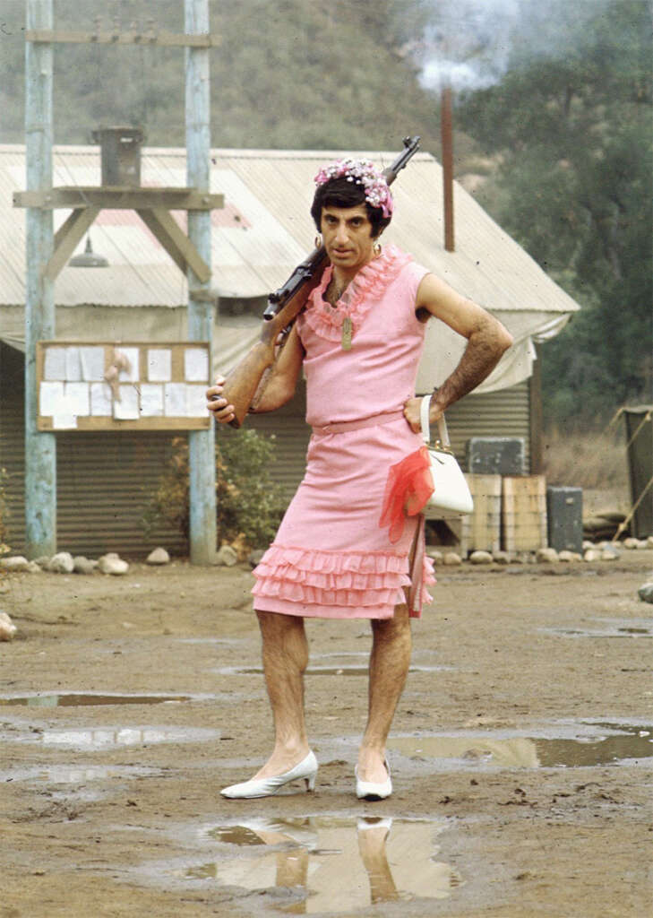 A person wearing a pink dress with ruffles and a floral headpiece stands holding a rifle on their shoulder. They have a pink purse, and are standing outdoors on a muddy surface with buildings in the background.