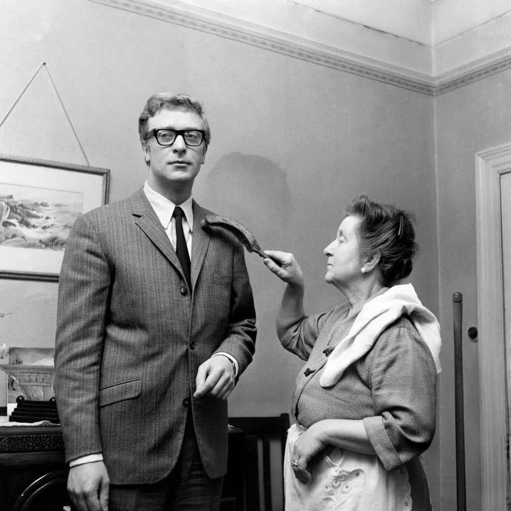 A woman in an apron brushes the shoulders of a man in a suit and glasses with a clothes brush. They are standing in a room with a picture on the wall behind them. The man looks off into the distance.