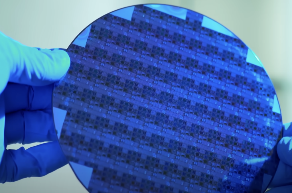 Gloved hands holding a blue silicon wafer with intricate circuit patterns, reflecting light.