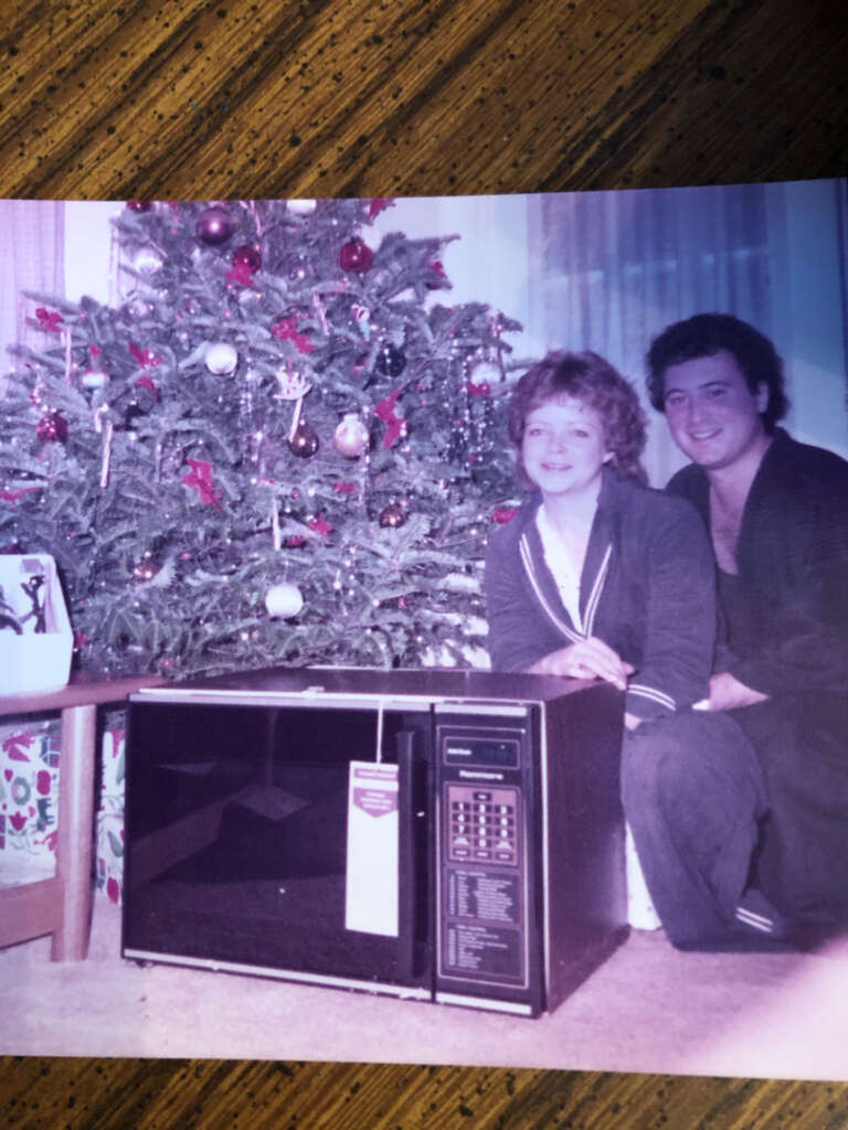 A smiling couple poses in front of a decorated Christmas tree with a new microwave oven on the floor. Wrapped gifts are under the tree, and the couple appears happy and relaxed in casual clothing.