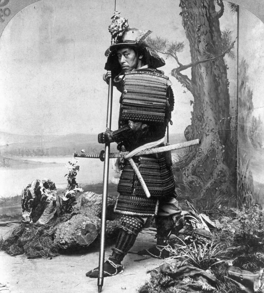 A historical photograph depicts a samurai in traditional armor holding a spear. The setting includes a painted backdrop of natural scenery with rocks, a tree, and rolling hills in the background. The samurai gazes directly at the camera.