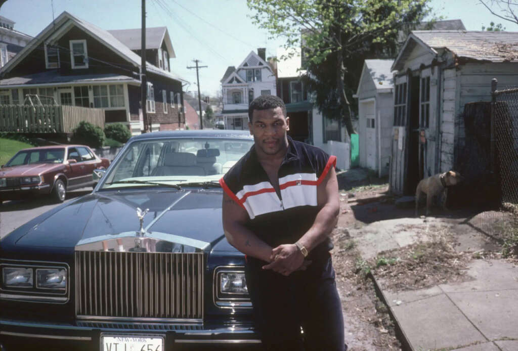 A man in a sleeveless shirt stands in front of a blue Rolls-Royce on a suburban street. There are houses and a dog in the background. The weather is clear and sunny.