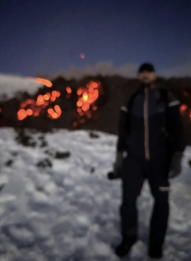 Blurry image of a person standing in snowy terrain during nighttime. In the background, glowing lava is visible, contrasting with the dark sky. The person appears to be wearing winter clothing and holding an object, possibly a camera.