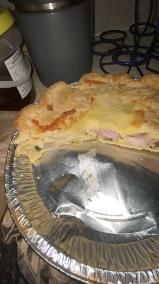 A partially eaten pie sits in a foil tray, revealing a filling of cream and ham chunks. The pastry is golden brown. In the background, there’s a cup and a jar on the left.