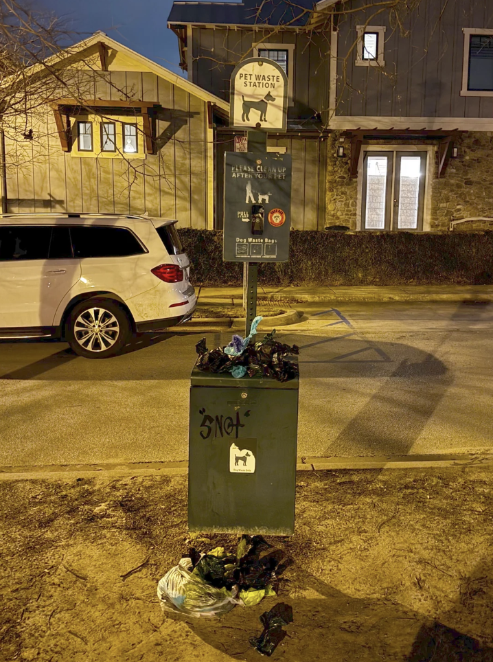 A pet waste station overflowing with plastic bags and waste at night. Bags are piled on the ground. Nearby is a parked white SUV and a house in the background. The scene is dimly lit by streetlights.