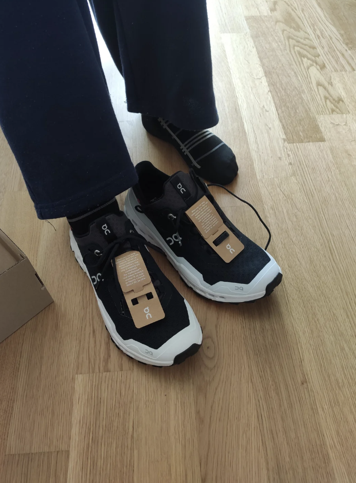 A person wearing black and white sneakers with tags still attached, standing on a wooden floor. One foot is partially out of the shoe, revealing a black sock. A bit of a cardboard box is visible in the corner.