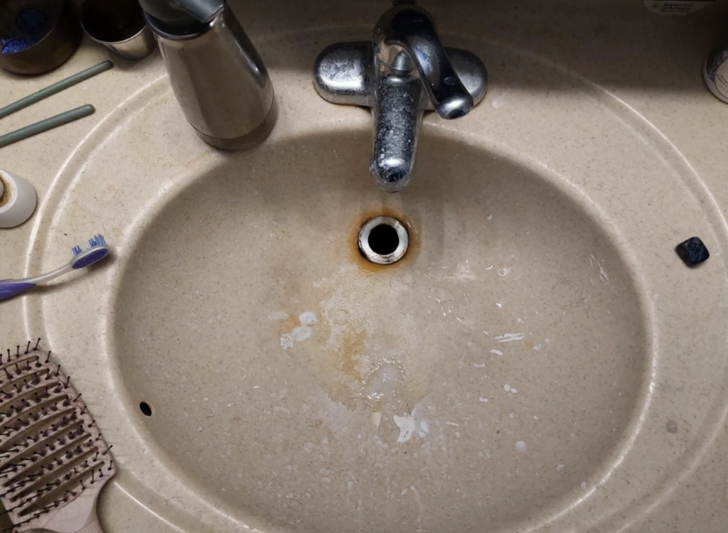 A bathroom sink with a beige countertop shows signs of rust around the drain. Watermarks and stains are visible on the surface. A toothbrush, hairbrush, and a soap dispenser are placed nearby.