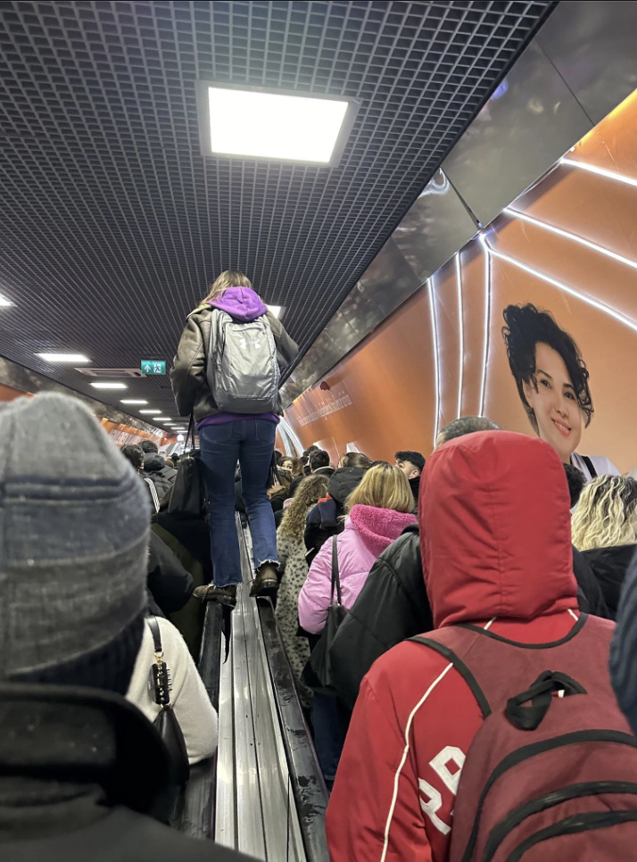 Crowded underground station with people standing on stairs and escalators. A person in a brown jacket and backpack stands atop the escalator divider. Bright orange walls feature an advertisement with a smiling face.