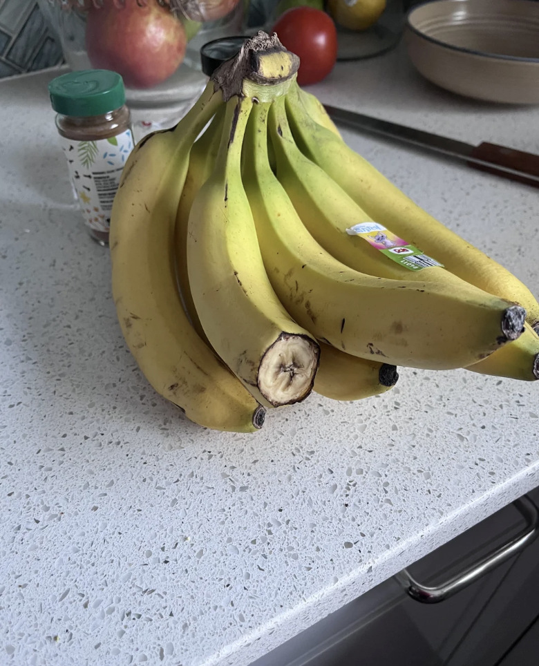 A ripe bunch of bananas rests on a white speckled countertop, next to a small spice jar and a kitchen knife. In the background, there is a bowl of apples and tomatoes.