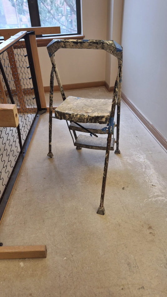 A worn, paint-splattered folding chair with a small platform stands on a scuffed floor near a window. Pieces of wood and a metal frame are visible nearby.