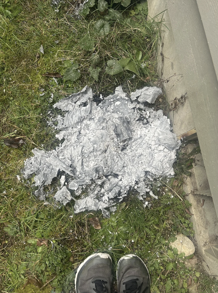 A pile of ashes lies on grass next to the base of a structure. Nearby, the tops of two sneakers are partially visible at the bottom of the image.