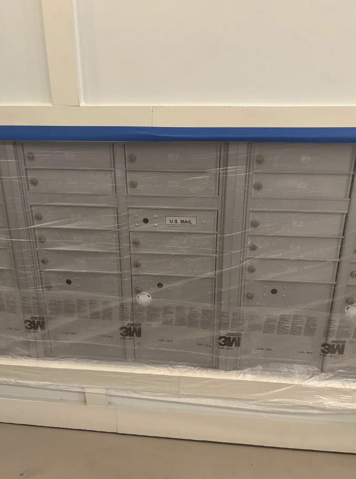 Gray metal mailboxes with various numbered compartments, from 161 to 166, are covered with transparent plastic wrap. A central compartment is labeled "U.S. MAIL." The bottom of the plastic sheet displays the "3M" logo.