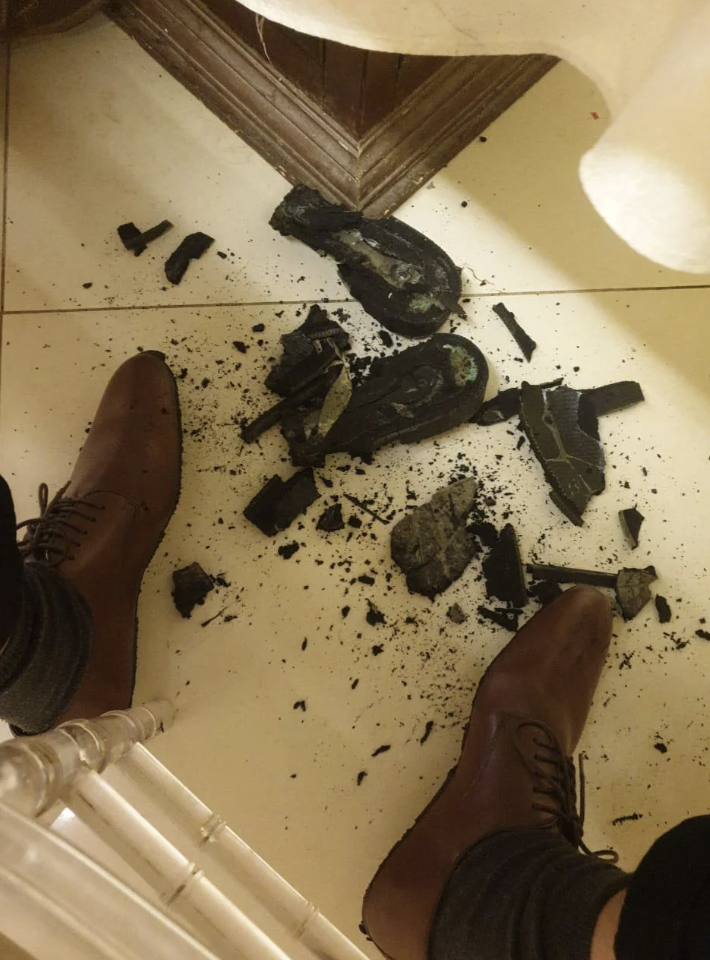 Brown shoes are seen on feet next to a broken or shattered dark object with scattered pieces on a light-colored tiled floor. The debris appears to include chunks and shards, possibly of a ceramic or glass item. A chair leg is visible on the left.