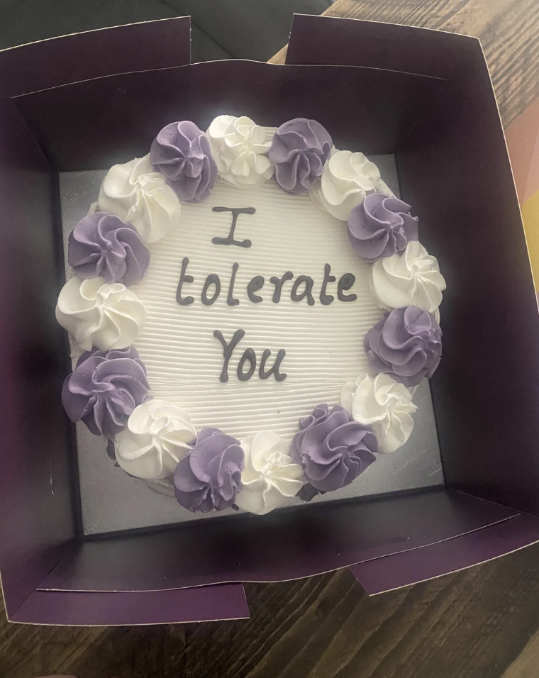 Round cake in a box with the words "I tolerate you" on top. The cake is decorated with alternating purple and white frosting rosettes around the edge.