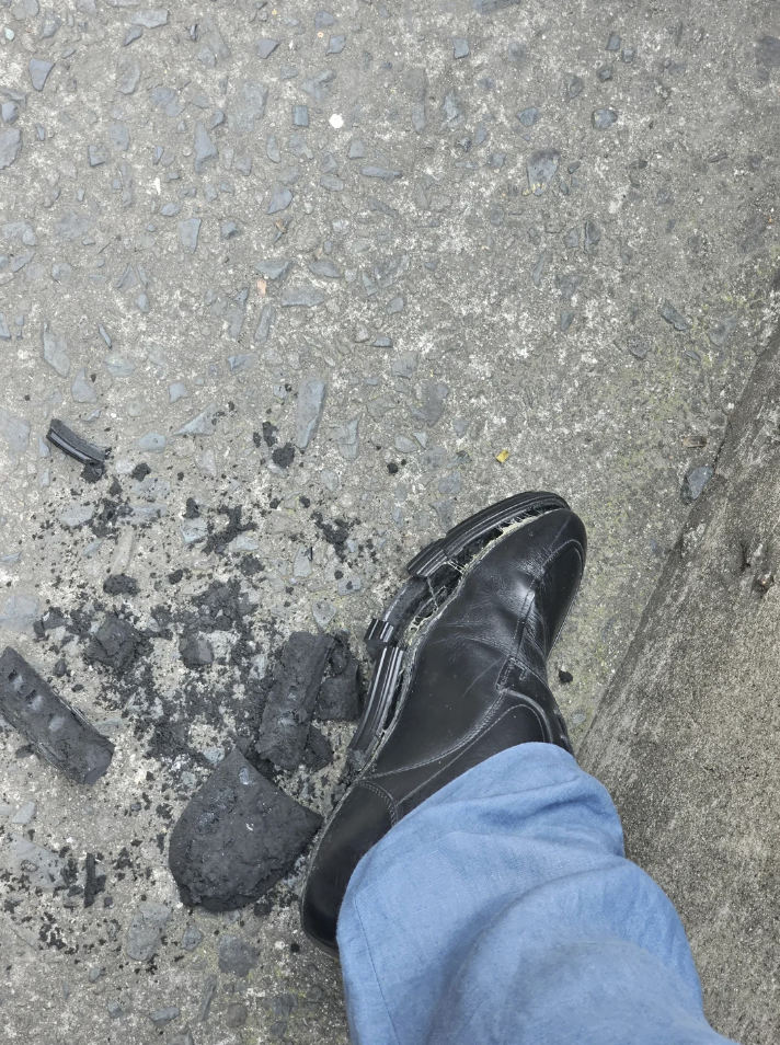 A worn black shoe with a damaged sole, surrounded by pieces of black debris, is on a textured gray pavement. The person is wearing light blue jeans.