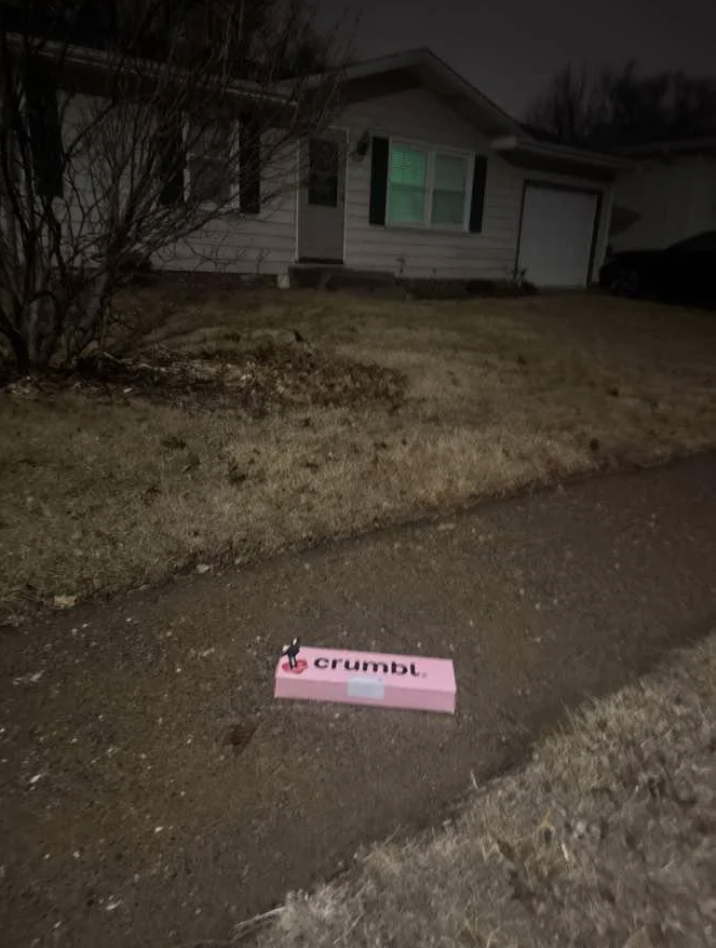 A pink Crumbl cookie box sits on a wet sidewalk in front of a house at night. The house has white siding and black shutters, with a dim light visible in one window. The surrounding yard is grassy and dimly lit.