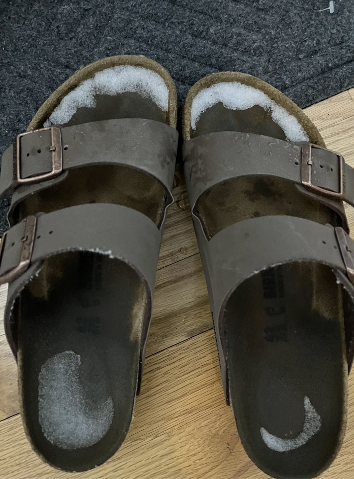 A pair of brown sandals on a wooden floor, each with snow or white frost collected at the toe and heel areas. The floor has a partial gray mat visible in the background.