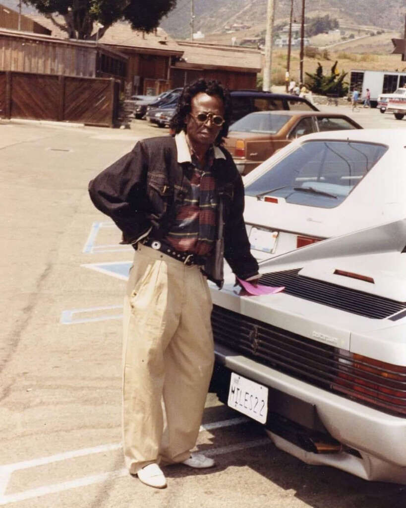 A person wearing sunglasses, a dark jacket, plaid shirt, and light-colored pants stands beside a silver sports car in a parking lot. The setting includes wooden buildings and hills in the background.