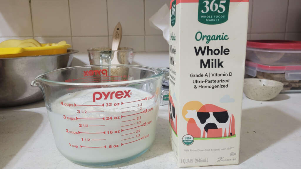 A Pyrex measuring cup filled with milk is next to a carton of 365 Organic Whole Milk on a kitchen counter. The cup is marked with measurements up to 4 cups. The carton displays a cow illustration and mentions it is ultra-pasteurized and homogenized.