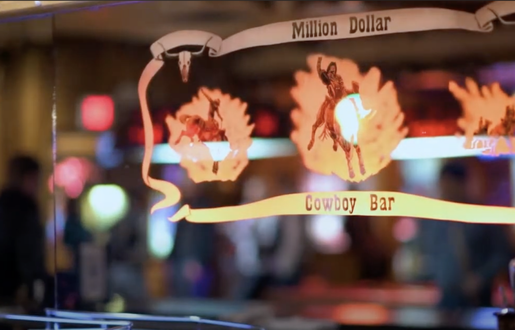 A neon sign for the "Million Dollar Cowboy Bar" is illuminated, featuring silhouettes of a bucking horse and rider. The background is blurred with various colored lights, suggesting a lively atmosphere.