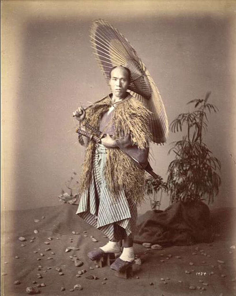 A man in traditional Japanese attire stands with a parasol and straw cloak. He wears sandals and holds a sword. The background features rocks and a potted plant, giving a natural setting indoors. The image is sepia-toned.