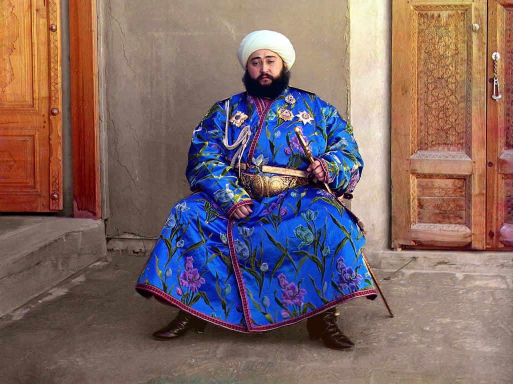 A man in a vibrant blue embroidered robe with floral patterns and a white turban sits against a wooden door backdrop. He wears a broad belt and black boots, exuding a regal presence.