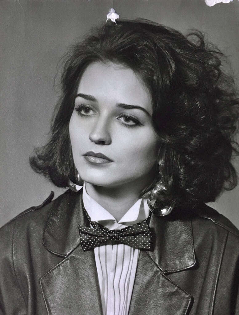 Black and white portrait of a woman with shoulder-length dark hair, wearing a leather jacket, a white pleated shirt, and a polka dot bow tie. Her expression is calm and contemplative.