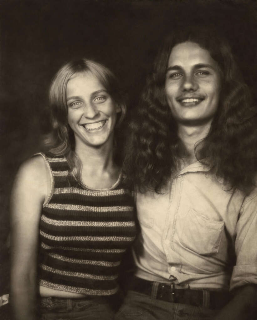 A black and white photo of two smiling people; one with long, straight hair and a striped tank top, the other with long, curly hair and a button-up shirt. They are sitting close together, both looking at the camera.