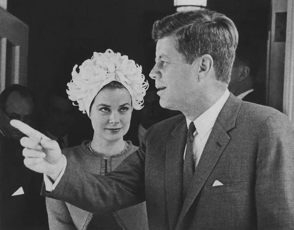 A black and white photo of a man in a suit gesturing while speaking, standing next to a woman in an elaborate hat and dress. They appear to be engaged in conversation outside a building.