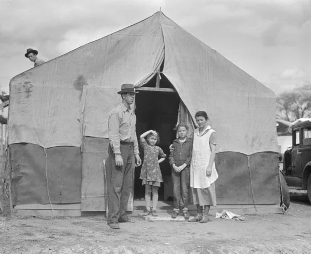 A black-and-white photo of a family standing in front of a large canvas tent. There are two adults and two young girls. A man is seen walking in the background. A vintage car is parked to the right of the tent. Cloudy sky overhead.