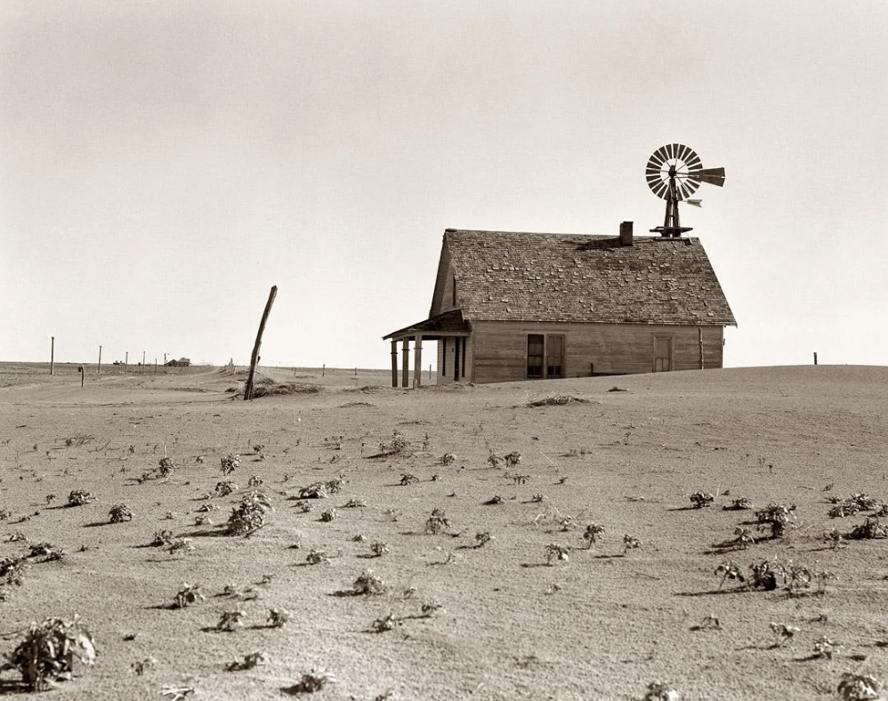 A lone, weathered wooden house with a windmill stands surrounded by a barren, dusty landscape. Sparse vegetation dots the dry ground under a clear sky, evoking a sense of isolation and desolation.