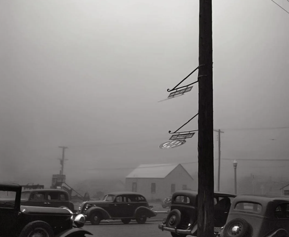 Vintage black and white photo of an early 20th-century street scene, featuring antique cars parked near a tall wooden pole with hanging signs. A hazy sky and classic architecture in the background suggest a nostalgic, foggy day.