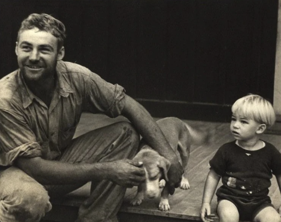 A man sitting on steps smiles while petting a dog. Next to him, a young child wearing a dark outfit with a belt stands, looking away. The background is a dark wall. The image has a vintage, sepia tone.