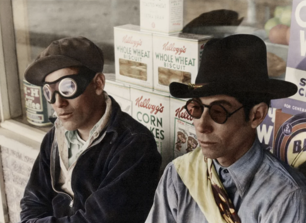 Two individuals wearing hats and goggles sit in front of a store window displaying vintage Kellogg's cereal boxes. One wears a dark jacket, the other a light shirt with a tie.