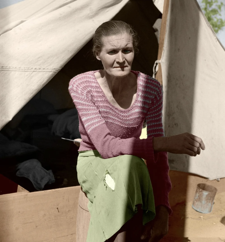 A woman sits outside a canvas tent, wearing a pink striped sweater and a green skirt. Her expression is contemplative, and she holds her hand on her knee. The tent is partially open, revealing a glimpse of the interior.