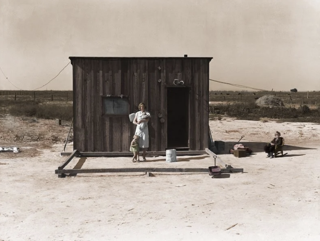 A woman stands holding a baby outside a small wooden shack in a barren landscape. A child stands beside her, holding her hand. A toddler sits nearby on a makeshift chair. The ground is sandy and the sky is overcast.