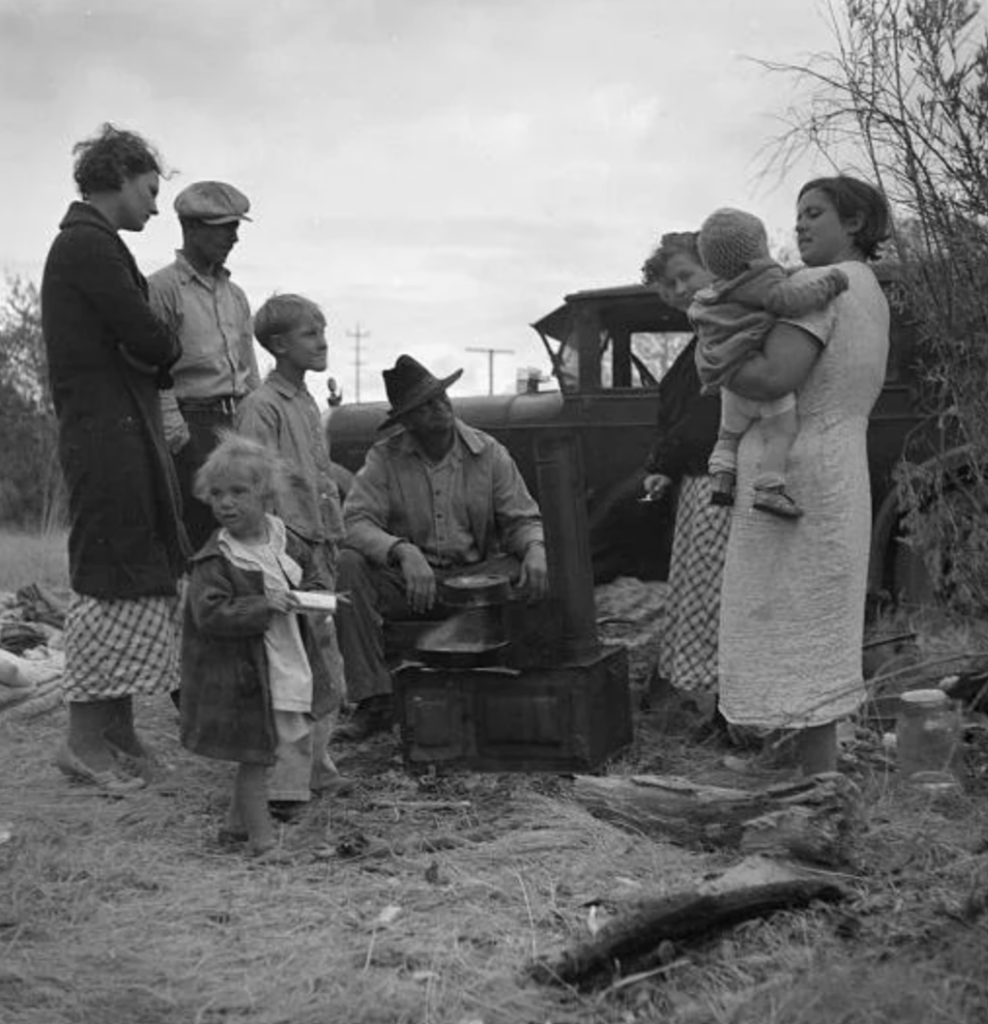 A black-and-white photo of a group of people gathered outdoors near a car. Adults and children stand around a seated person cooking on a small stove. The scene has a rustic feel, with dry grass and sparse trees in the background.