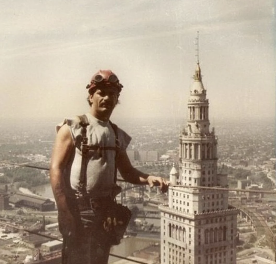 A construction worker in a sleeveless shirt and safety gear stands on a high ledge, with a panoramic cityscape in the background. A tall, ornate building with a spire is prominently visible beside him.